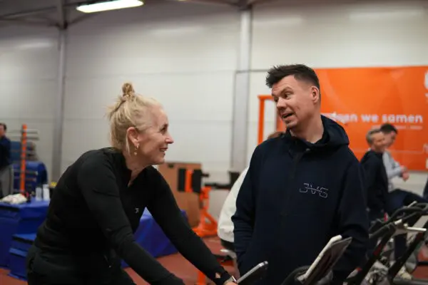Een vrouw traint onder begeleiding van Jelle van Gorkom op een hometrainer in een fitnesszaal.
