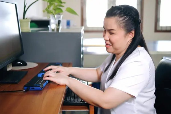 Vrouw werkt achter computer met brailletoetsenbord op bureau in lichte kantoorruimte.