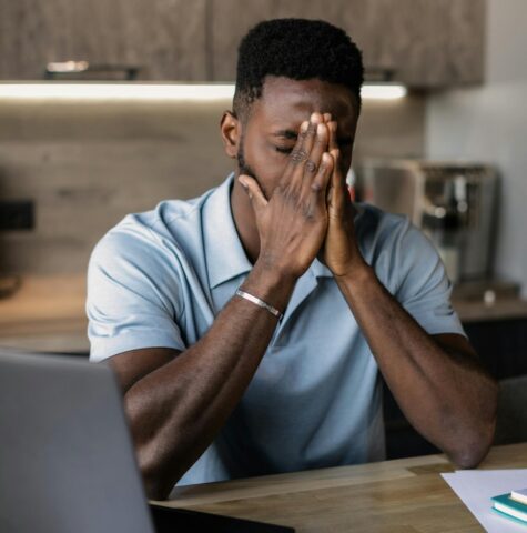 Man zit met handen voor gezicht achter laptop aan tafel, oogt vermoeid of gestrest tijdens het werken.