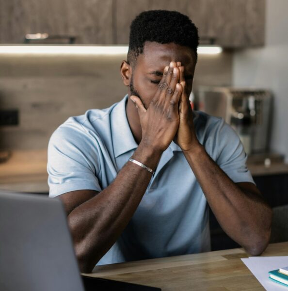 Man zit met handen voor gezicht achter laptop aan tafel, oogt vermoeid of gestrest tijdens het werken.